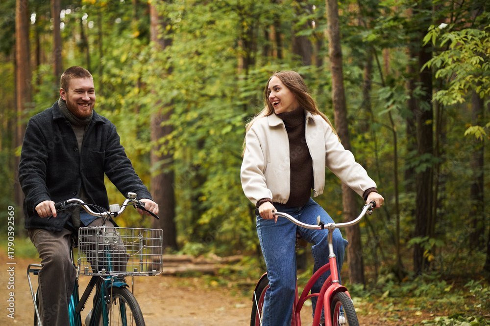 Obraz premium Caucasian young man and Caucasian young woman riding bicycles through forest path, smiling and looking at each other, surrounded by green trees, enjoying outdoor activity together