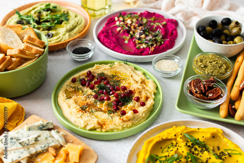 A vibrant spread showcases different types of hummus including classic, beetroot, broccoli, and turmeric. Accompanied by olives and breadsticks, it invites sharing and enjoyment