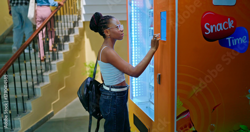 African young adult gen z lady woman use easy fast self service juice drink press button campus place. Happy pupil girl wear bag enjoy break time day indoor college hallway area 