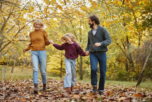 A family of three walks hand in hand through a vibrant park filled with colorful autumn leaves. They smile and share joyful moments on a sunny weekend.