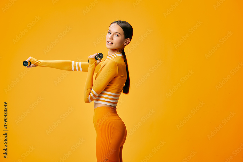 Fototapeta premium Woman in bright orange athletic wear holding dumbbells against a vibrant orange backdrop, showcasing fitness training, balance, and athletic confidence.