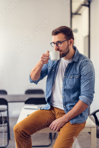 Young office worker drinking coffee on break and relaxing leaning on office desk.