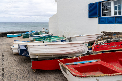 Εκτύπωση καμβά A collection of colorful rowboats sit on the shore near a white building with blue accents, by the calm ocean