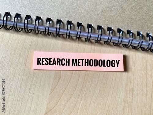 Word Research Methodology on a sticky note strip below the spiral binding of a notebook. Represents the systematic process, design, and techniques used to conduct academic or scientific research.