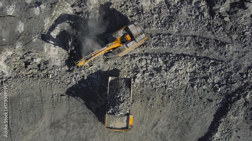 Aerial top-down view of an excavator loading rocks into a dump truck at an open-pit mine. Industrial drone footage showing heavy machinery working in a dusty environment, highlighting extraction