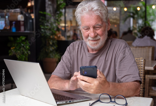 Photos Smiling bearded senior man sits in a cafe using laptop and smartphone