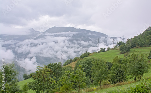  Green meadow with mountain peaks covered in mist and clouds. La Vanoise national park, Savoie, France 