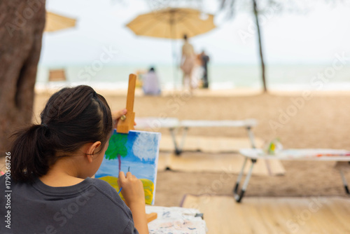 Over-the-shoulder view with blurred background of a young girl painting a seascape on an easel at beautiful beach, representing childhood creativity, outdoor learning, and a peaceful artist hobby.