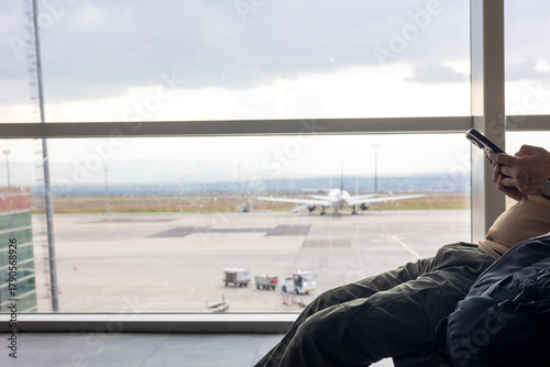 Passenger relaxes and uses a smartphone while waiting for a flight in the airport terminal. The view through the large window shows an airplane, evoking a sense of a calm journey and modern travel.