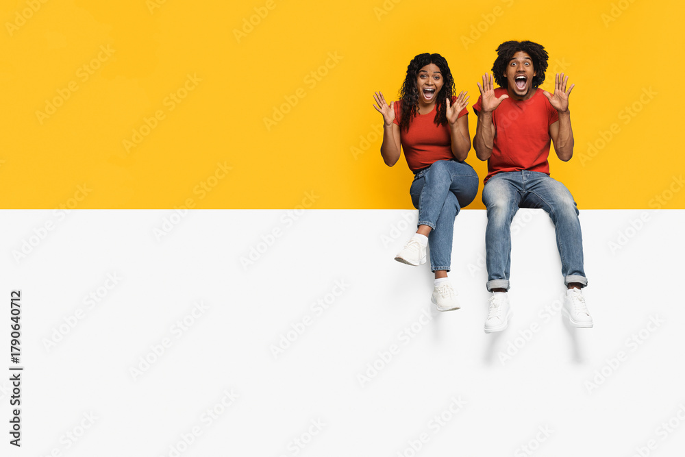 Fototapeta premium A happy couple shows excitement by sitting on a white surface. They wear matching red shirts and jeans, with big smiles and arms raised in celebration against a vibrant yellow background.