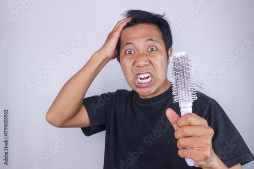 Carta da parati funny expression of angry asian man holding comb, hair loss