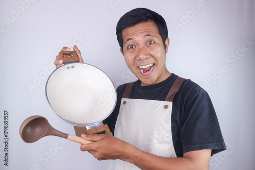 Happy expression asian man chef wearing apron holding pan and soup spoon on white background