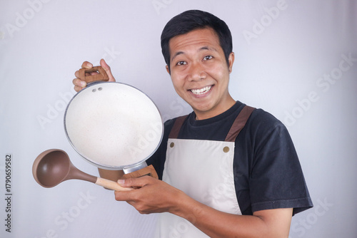 Smiling face expression asian man chef wearing apron holding pan and soup spoon on white background