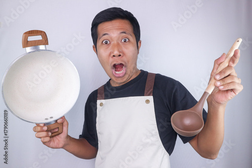 Funny excited expression asian man chef wearing apron holding pan and soup spoon on white background