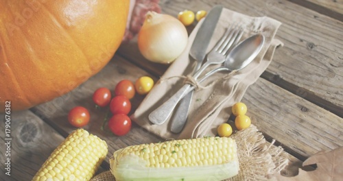 Showing partly husked corn resting on wooden plank, with pumpkin, tomatoes, onion and tied cutlery