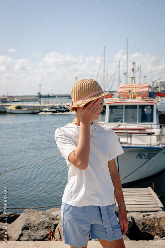 Fototapeta premium Girl standing on a pier near boats and yachts, wearing a hat and summer outfit. Relaxed vacation atmosphere by the marina under a bright blue sky. 