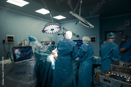 In a brightly lit Hospital operating room, a surgical team wearing gowns and masks performs a knee replacement with robotical surgery robotic assistance while monitoring progress on screens