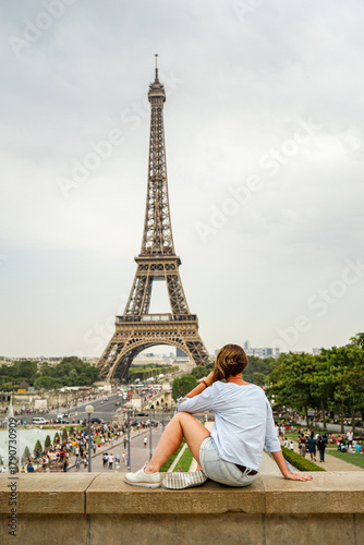 A girl in a blue blouse, with a short hairstyle, wearing shorts and white shoes, is sitting on a rock with the Eiffel Tower in the background in Paris, France The weather is cloudy in the evening