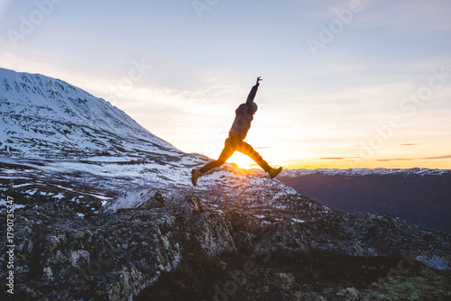 Traveler jumps from rocks at sunset near Gaustatoppen Mountain in Norway. Golden light, snow peaks, and open sky capture pure joy and freedom of adventure