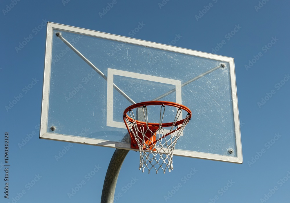 Naklejka premium A clear acrylic basketball backboard mounted high above an outdoor court, featuring a vibrant orange rim and white nylon net ready for game play ,sport ,practice ,durable