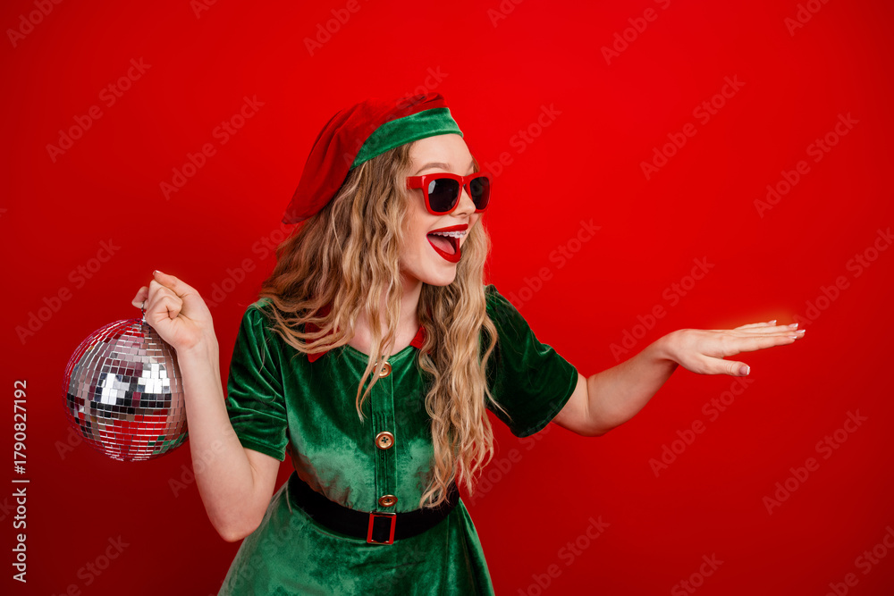 Naklejka premium Festive young woman dressed as an elf holding a shiny disco ball and striking a playful pose with excitement