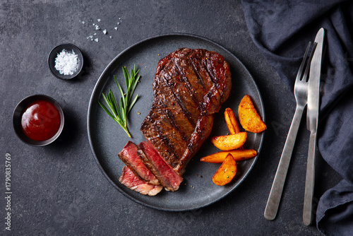 Beef Steak medium rare with roasted potato wedges on a black plate. Grey background. Close up. Top view.