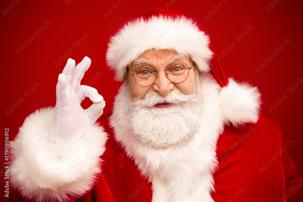 Naklejka premium Santa in classic red suit with fluffy white trim and round glasses gives an ok sign against a red background perfect for christmas season