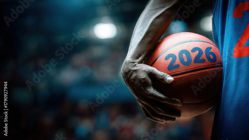 Athlete in blue jersey holds basketball with «2026» inscription, stadium lights create bokeh effect in background. Vibrant sports event atmosphere