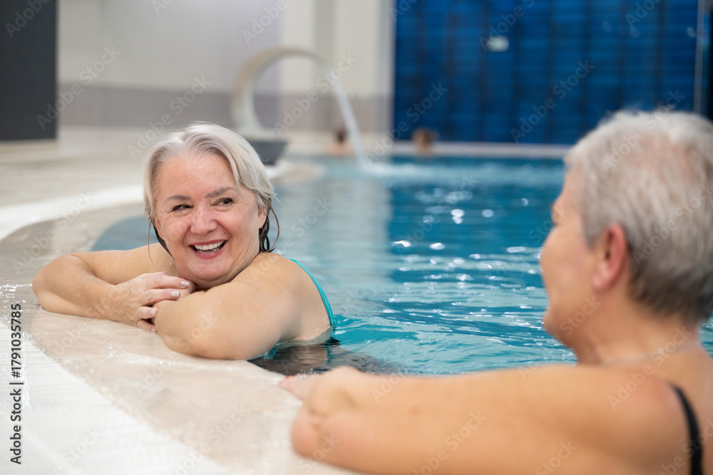 Fototapeta premium Two smiling senior women enjoying leisure time in an indoor swimming pool, leaning on the poolside. Concept of friendship, wellness, active lifestyle, relaxation, and healthy aging.