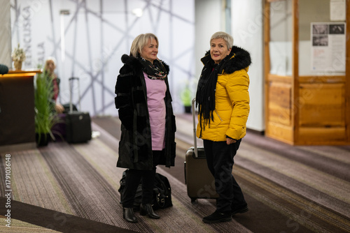 Two older women dressed in winter clothing standing in a hotel lobby and having a friendly conversation next to their luggage. Concept of travel, friendship, and leisure lifestyle for seniors.