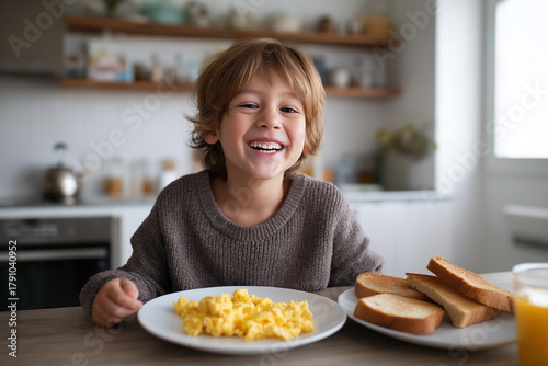 Happy young boy smiling with delight at breakfast, with scrambled eggs and toast on the table. Sunny morning meal for a child.