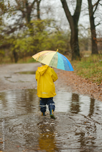 Happy child in yellow raincoat and hat holding colorful rainbow umbrella standing in puddle on rainy autumn day in park.