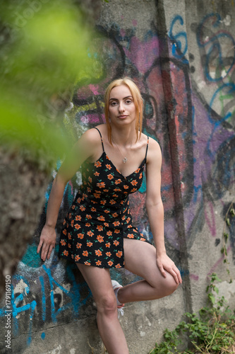 Blonde sensual woman with long legs posing confidently in front of a colorful graffiti wall, wearing a short floral dress. Urban fashion meets vibrant street art in this expressive portrait.