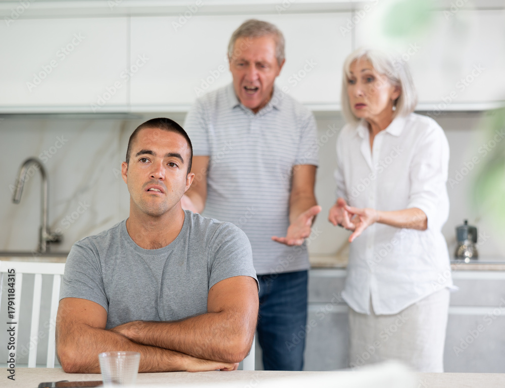 Fototapeta premium Family of threen - mother, father and son quarrel in kitchen. Eldery cuple is disappointent of adult son.