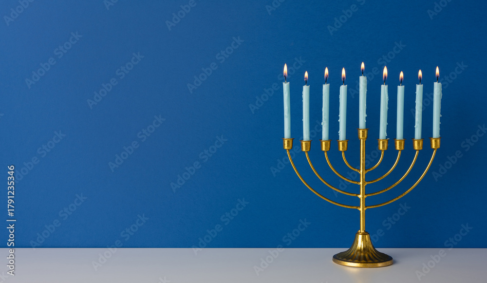 Obraz premium A golden menorah with nine lit candles sits on a white surface before a deep blue wall, evoking Hanukkah tradition, warmth, and holiday celebration