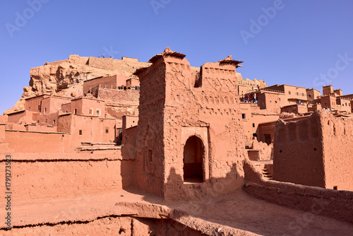 Structures made of earthen materials stand under a cloudless sky in Ait Ben Haddou. Ancient architecture and desert landscape are seen in Morocco.