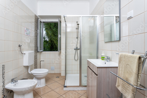 A contemporary bathroom showcases a walk-in shower, toilet, and bidet, with large tiles and natural light streaming through the open window