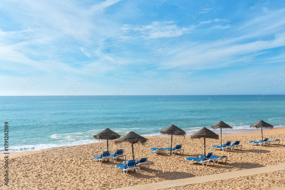 Fototapeta premium Sun-kissed sandy beach features lounge chairs arranged beneath thatched umbrellas, inviting relaxation by the calm ocean waves and clear blue skies