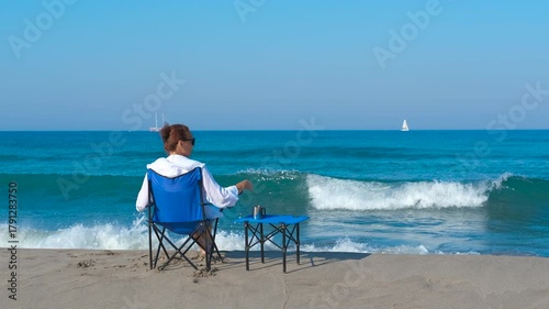 Woman relaxing on beach chair watching ocean waves. Successful woman resting on beach chair, gazing at sparkling ocean waves and distant sailboat, tranquil seaside freedom