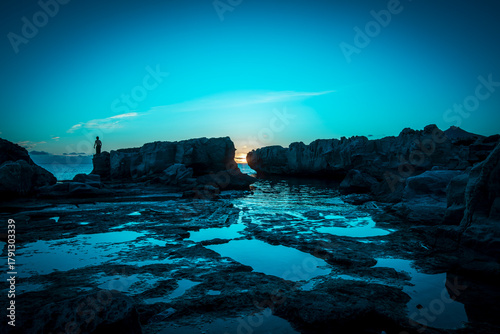 At sunset, a man stands ready to dive, waiting for the right wave on a rock on the island of Favignana, province of Trapani, Sicily, Italy.
