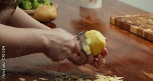 Close up of hands peeling fresh potato with peeler on wooden table, preparing ingredients for homemade vegan meal, cooking process, healthy food, rustic kitchen atmosphere, natural lifestyle