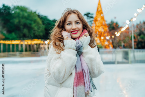 Portrait of happy beautiful girl wrapping in warm scarf trying to keep warm on background of outdoor ice skating rink. Winter holidays mood. Outdoor activities on Xmas and New Year.