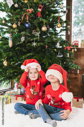 Two smiling kids in festive ugly sweaters and oversized Santa hats sitting under Christmas tree at home during winter holidays. Happy siblings feeling excited, having fun, waiting for Christmas gifts.