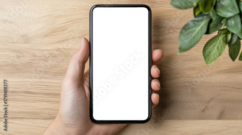 hand holding smartphone with blank white screen on wooden desk, perfect for mockup, app design, and modern presentation visuals