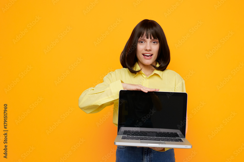 Naklejka premium Cheerful young woman holds a laptop, presenting a bright digital scene. She smiles, wearing a yellow shirt, standing against a bold orange background.