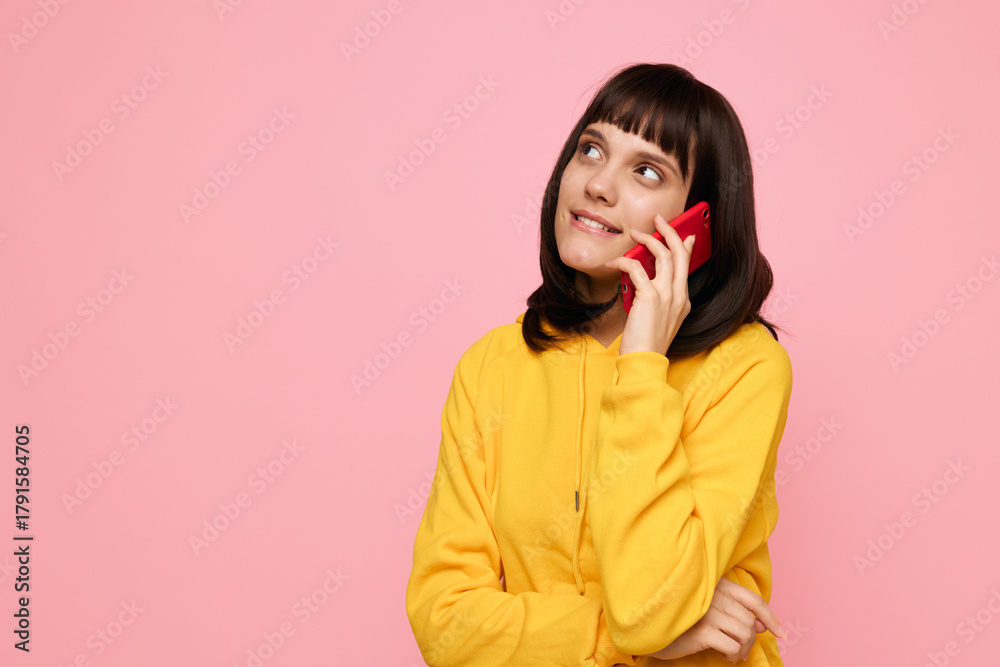 Fototapeta premium A young woman with dark hair and bangs wears a bright yellow hoodie and talks on a red mobile phone against a soft pink background, conveying a cheerful casual mood.