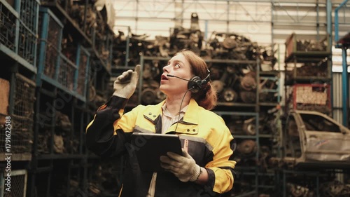 One Hispanic mature female inspector remotely works with headset and tablet, checks auto spare parts stock in warehouse stack, operator jobs in manufacturing factory, and industrial worker occupation.