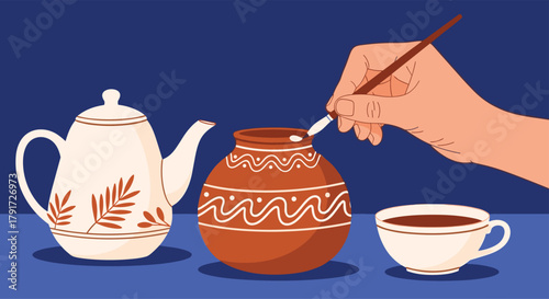 A hand decorating a clay pot with white paint on a blue surface next to a teapot and a cup of tea.