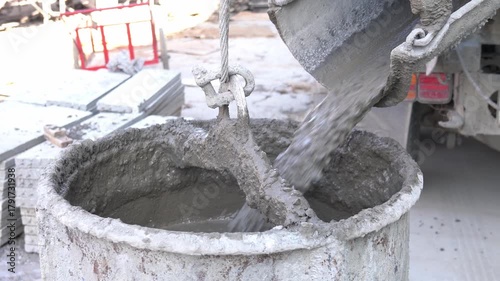 A close-up mixed wet concrete being poured from the chute of a concrete mixer truck into a large, sturdy bucket suspended by a wire cable.