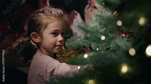 A young girl decorates a Christmas tree with glowing lights, surrounded by festive decorations and family members preparing for the holidays at home
